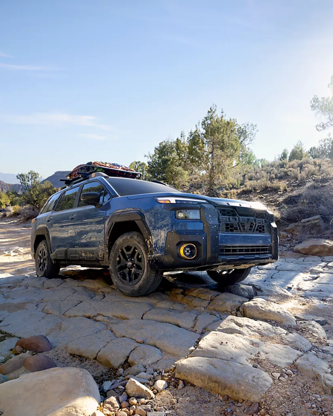 2026 Subaru Outback driving on a rocky terrain. The SUV is dark blue and there are trees in the background.