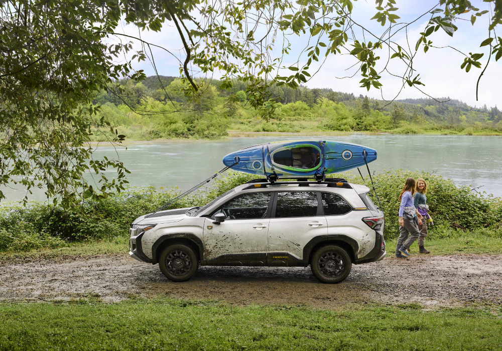 Driver's side exterior view of s 2026 Subaru Forester Wilderness parked in front of a large body of water. There is a kayak strapped to the roof rack, and two women are walking away from the car
